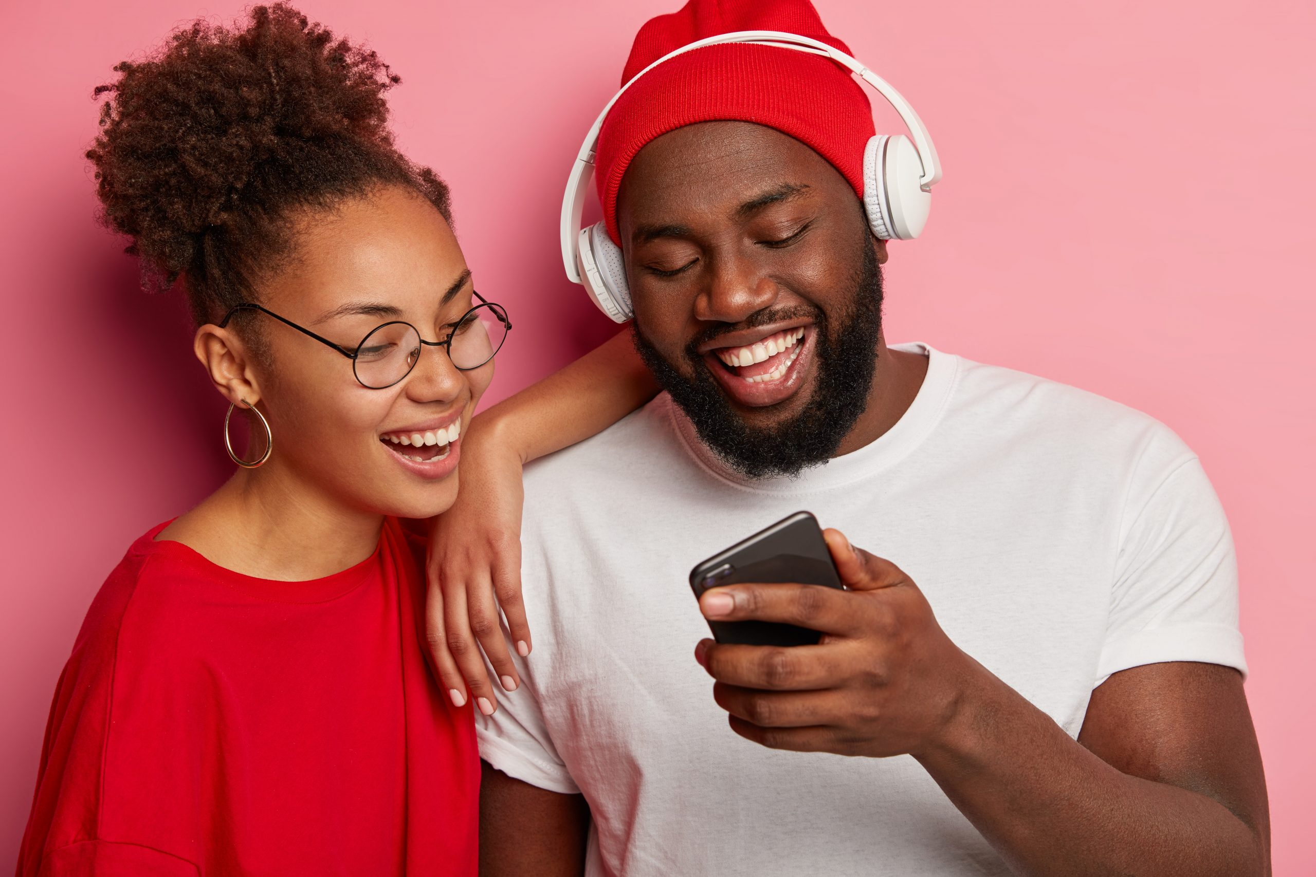 Happy ethnic woman and man watch funny video on smartphone, black man in red hat and white t shirt, wears headphones, shows new app to girlfriend, have fun together, pose over pink studio wall