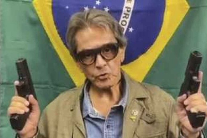 1_roberto_jefferson_segurando_duas_armas_uma_em_cada_mao_na_frente_de_uma_bandeira_do_brasil_1_38004-26144966