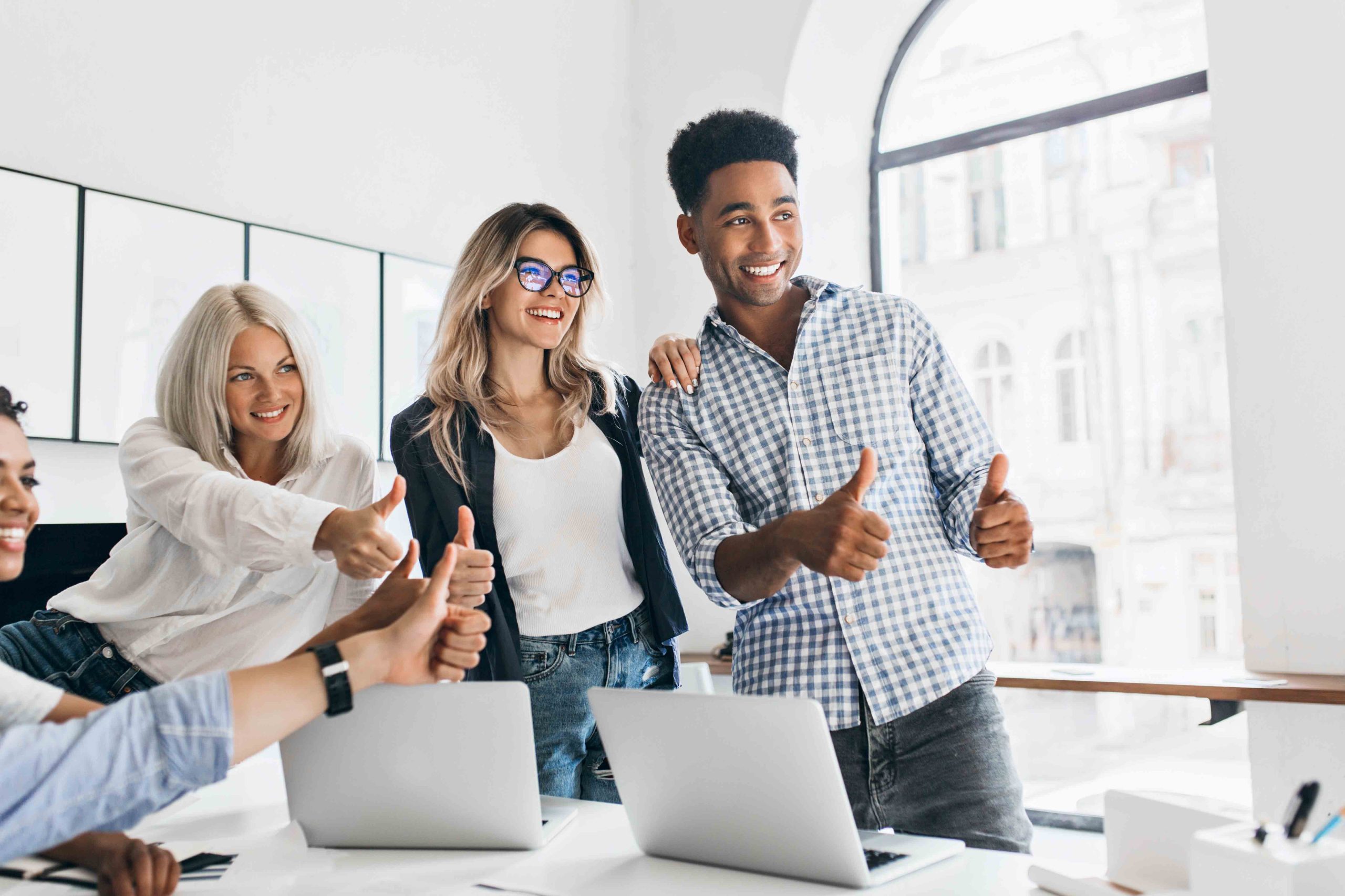 Tall african manager with big smile holding thumbs up and looking away. Charming girls working as managers posing in office next to table with laptops on it and laughing..